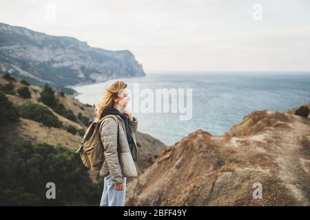 Happy young traveler woman walking and enjoying amazing coastline mountain view. With beige backpack, wearing in jacket and jeans. Stock Photo