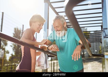Checking training results. Happy mature family couple in sports clothing looking at smartwatch and smiling while exercising together outdoors, they Stock Photo