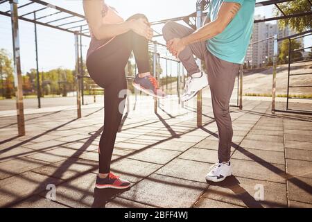 Stretching exercises. Cropped photo of man and woman in sportswear warming up together while standing at the stadium. They are stretching legs. Active Stock Photo