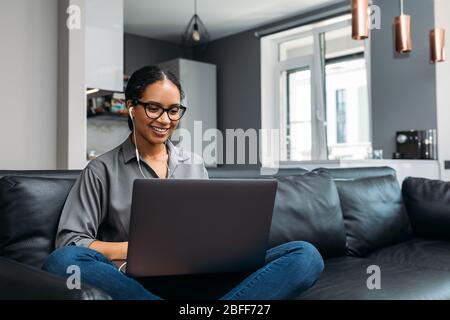 Young woman video calling using a laptop sitting on a sofa wearing earphones Stock Photo