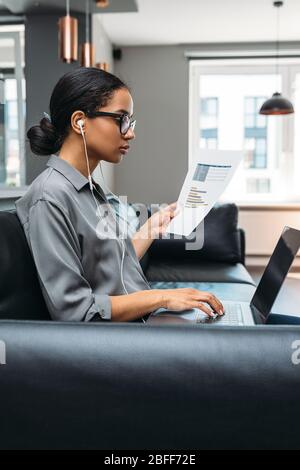 Young businesswoman working from home. Side view of freelancer using laptop computer sitting on the sofa wearing eyeglasses. Stock Photo