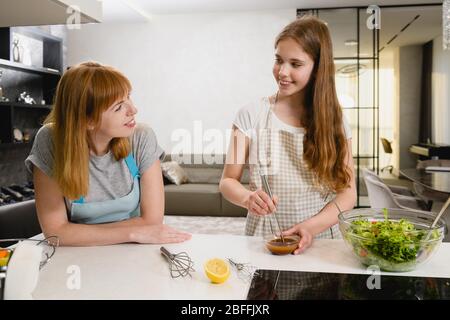 Two smiling women cut green salad with tomatoes and mix dressing in small bowl with lemon juice, dark sauce and oil at kitchen Stock Photo