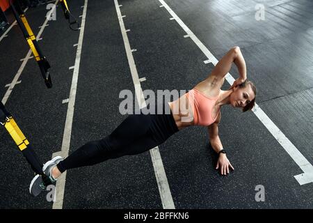 Sporty Lifestyle. Young woman doing side-plank tap TRX exercise at gym smiling confident Stock Photo