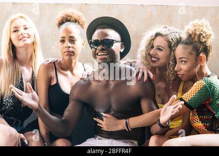 Young black man with sunglasses and hat gesturing with hands surrounded by girls sitting on a sofa Stock Photo