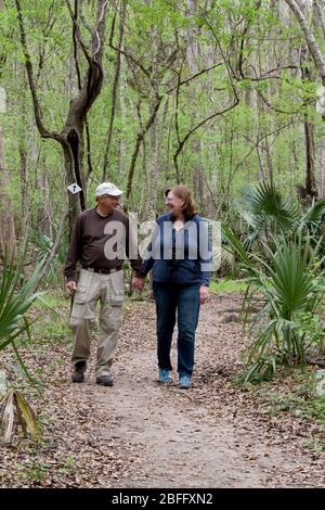 An active retired senior couple walking a trail in a forest with trees and palms looking at each other Stock Photo