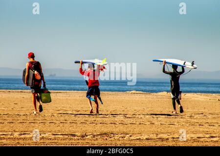 Hefting their boards and wearing wet suits, three surfers head for the waves in Huntington Beach, CA. Stock Photo