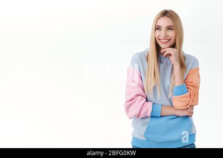 Portrait of attractive blond young woman in hoodie, looking left and laughing with beaming pleased smile, reading funny promo on white empty space for Stock Photo