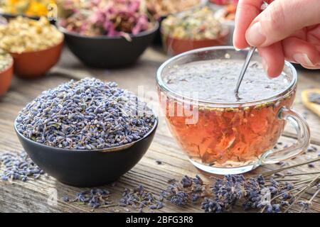Healthy lavender tea cup being stirred by a woman hand, on a wooden table top surrounded by bowls of dry medicinal herbs. Herbal medicine. Stock Photo