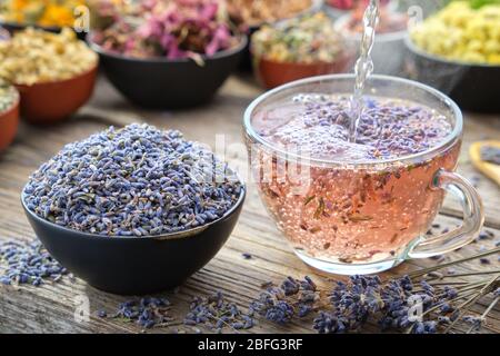 Healthy lavender tea cup being stirred by a woman hand, on a wooden table top surrounded by bowls of dry medicinal herbs. Herbal medicine. Stock Photo
