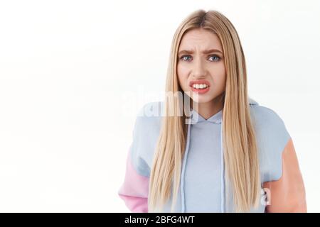 Close-up portrait of embarrassed blonde girl, stooping and clench fists, grimacing as staring something cringy, standing in awkward awful situation Stock Photo