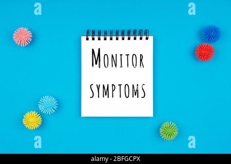 Paper notebook with plastic balls as viruses on the blue background with Monitor your symptoms wording. Epidemic, social isolation, coronavirus COVID-19 concept. Isolated white paper Stock Photo