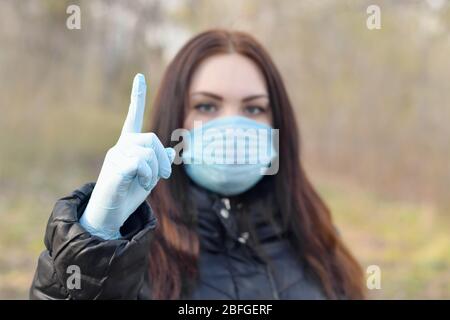 Portrait of young brunette woman in blue protective mask and rubber gloves shows attention gesture outdoors in spring wood. Concept of protective good Stock Photo