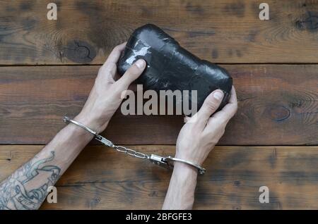 Arrested drug dealer hands in police handcuffs with big heroin drug package on dark wooden table background. Illegal drug trafficking concept. Fightin Stock Photo