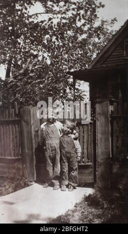 1930s black and white photo of two brothers wearing denim overalls standing in front of a wooden fence. Stock Photo