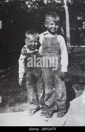 Close up of two brothers wearing denim overalls photographed in the 1930s with the big brother's arm around the little brother's shoulders. Stock Photo