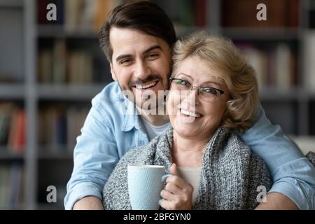 Elderly mother holding cup tea enjoys adult son warm embracement Stock Photo
