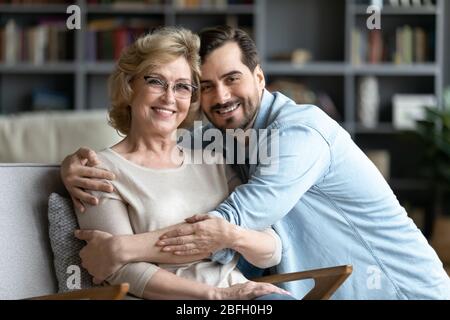 Adult son hugs elderly mother while she resting on armchair Stock Photo