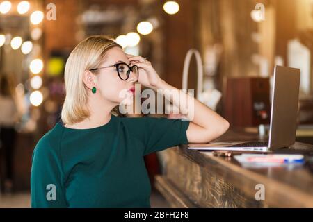 Portrait of beautiful blonde woman with short haircut in glasses looking with tired expression at laptop computer screen, holding head. Female adminis Stock Photo