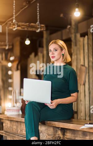 Cheerful elegant blonde business woman, administrator wearing emerald color suit sitting on wooden reception desk holding laptop, having fun in workpl Stock Photo
