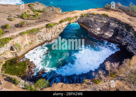 Aerial view of ocean waves crashing through a rocky archway into a round, isolated bay (Broken Beach, Nusa Penida, Indonesia) Stock Photo