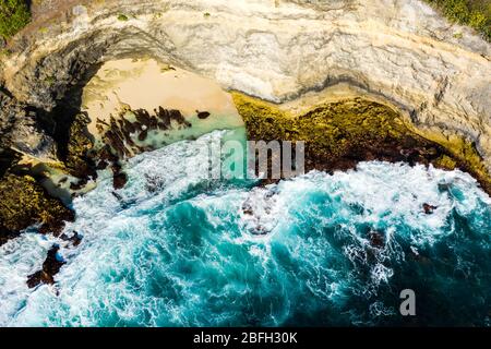 Top down aerial view of ocean waves crashing onto a small sandy beach surrounded by cliffs Stock Photo