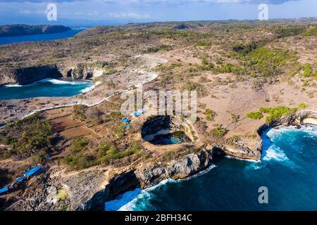 Aerial view of ocean waves crashing through a rocky archway into a round, isolated bay (Broken Beach, Nusa Penida, Indonesia) Stock Photo