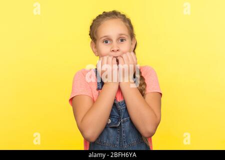 Portrait of frightened nervous little girl in denim overalls biting nails and looking at camera with scared big eyes, feeling worried, anxious about c Stock Photo