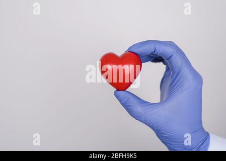 Cropped close up photo of hand in nitrile latex glove holding showing little red heart isolated over gray background copyspace Stock Photo
