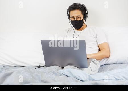 Man with protective mask and headphone working on laptop in bed at home. Stock Photo