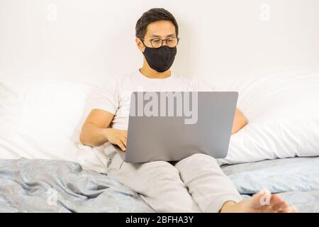 Man with protective mask working on laptop in bed at home covid-19 outbreak. Stock Photo