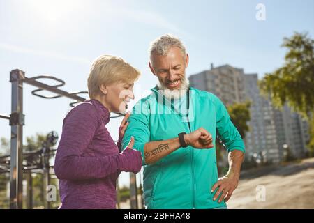 Great results. Happy mature family couple in sportswear looking at smartwatch and checking training results while standing outdoors Stock Photo