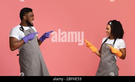 Cheerful African Couple In Aprons And Rubber Gloves Pointing At Each Other Stock Photo