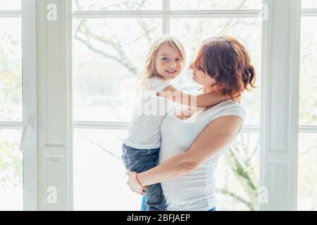 Stay Home Stay Safe. Young mother holding her child. Woman and little girl relaxing in white bedroom near windiow indoors. Happy family at home. Young mom playing with her daughter Stock Photo