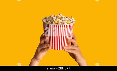 Unrecognizable man holding striped paper bucket with popcorn on orange background, close up. Panorama Stock Photo