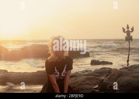 Trident totem near Beautiful young white skined woman sittind on Shiva Face Rock Carving on Vagator Beach, Goa, India. sunset background. Northernmost Stock Photo