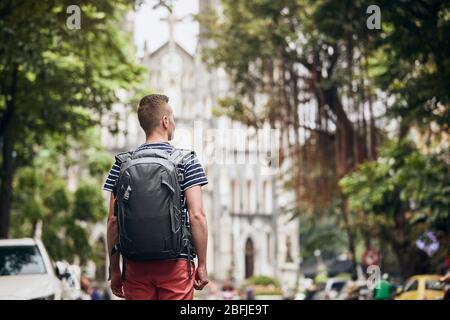 Traveler walking on street against cathedral. Rear view of young man with backpack in Hanoi, Vietnam. Stock Photo