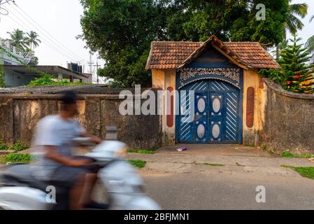 Kochi, Kerala, India - April 4, 2018: An unrecognisable man riding a scooter, blurred by motion, is passing in front of an ornate garden gate in the h Stock Photo