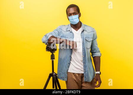 Portrait of cheerful photographer or video blogger with surgical medical mask smiling at camera with professional digital dslr camera on tripod. indoo Stock Photo