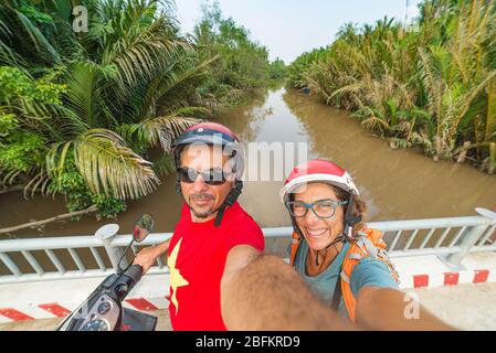Couple taking selfie on motorbike. Man and woman with helmet biking in the Mekong Delta region, South Vietnam. Lush green coconut palm tree woodland a Stock Photo