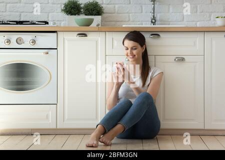 Smiling attractive young woman using smartphone sitting on kitchen floor. Stock Photo