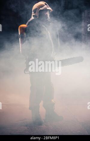 Portrait of young fireman standing and holding a chainsaw in the middle of the chainsaw's smoke Stock Photo