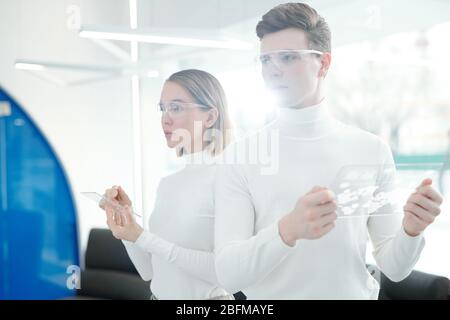 Two young contemporary progressive software developers in safety goggles and white jumpers using futuristic mobile gadgets during work Stock Photo