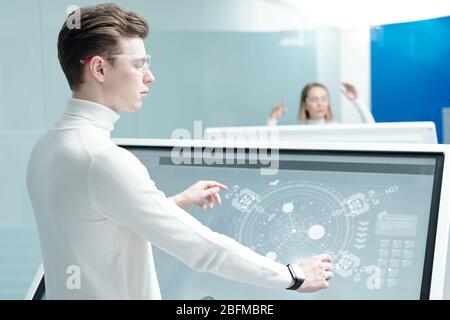 Young software engineer in smart goggles standing by large interactive display with holographic schemes while analyzing virtual data Stock Photo