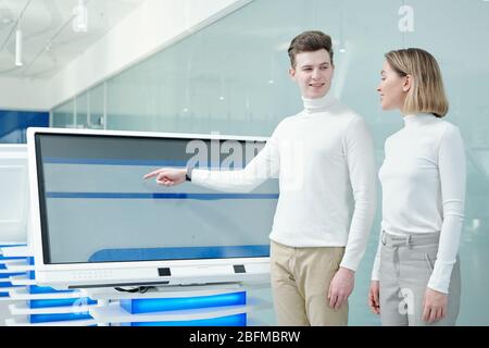 Happy young intelligent man looking at his colleague or customer while presenting new futuristic software on display in laboratory Stock Photo