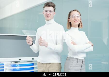 Two young smiling programmers in smart casualwear and goggles standing on background of large interactive display in office Stock Photo