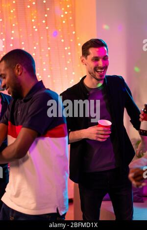 Laughing guy with red glass dancing and enjoying home party with friends with African man in t-shirt having fun near by Stock Photo