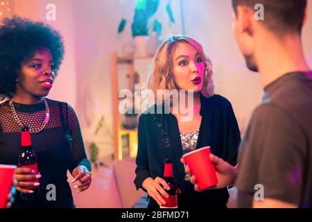 Young blond pretty woman with bottle of beer talking to her boyfriend at party while having drinks and fun with friends Stock Photo