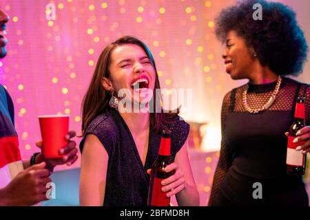Young laughing brunette woman with bottle of beer enjoying home party among her African friends on sparkling background Stock Photo