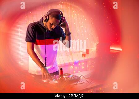 Young deejay of African ethnicity in headphones bending over desk with turntables while mixing sounds and making new music Stock Photo