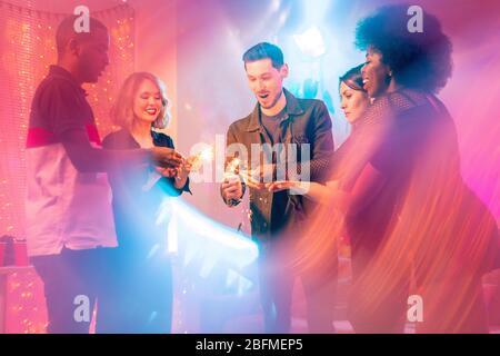 Five young intercultural friends standing in circle and holding sparkling bengal lights in the center while enjoying home party Stock Photo